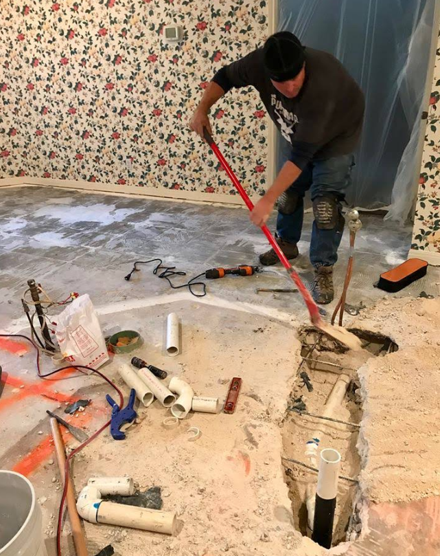 A worker uses a red pry bar to clear debris from a trench cut into a concrete floor, with plumbing pipes scattered nearby.