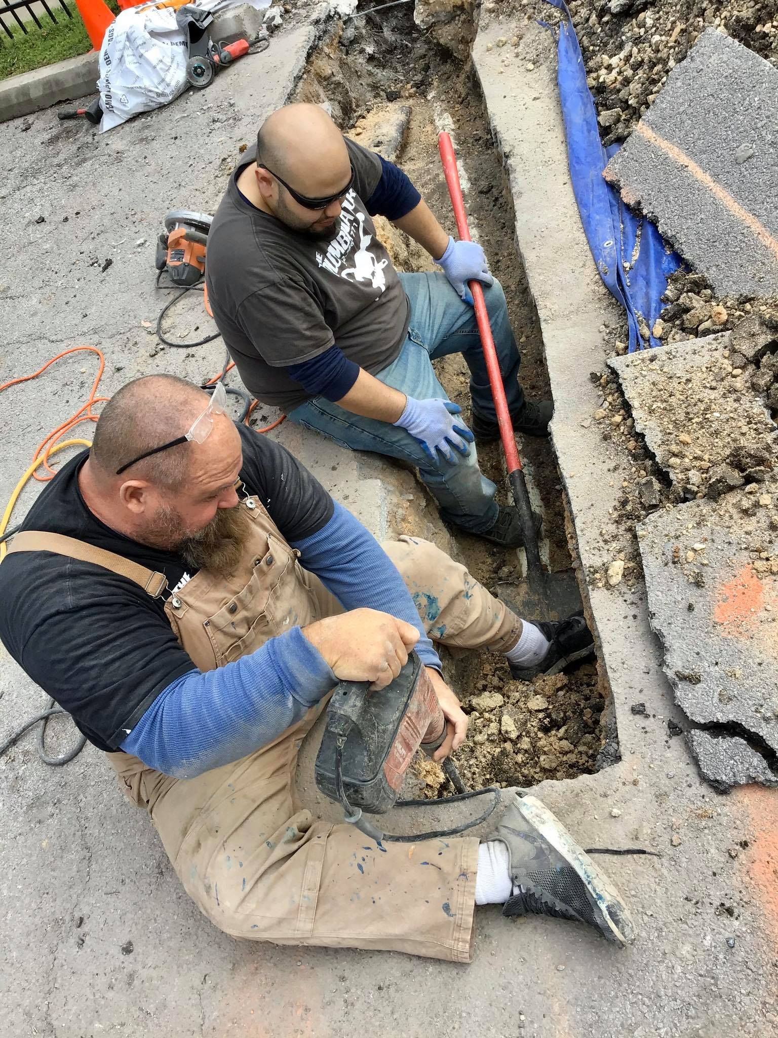 Two workers in work clothes and gloves use a shovel to clear dirt from a narrow trench along a road.