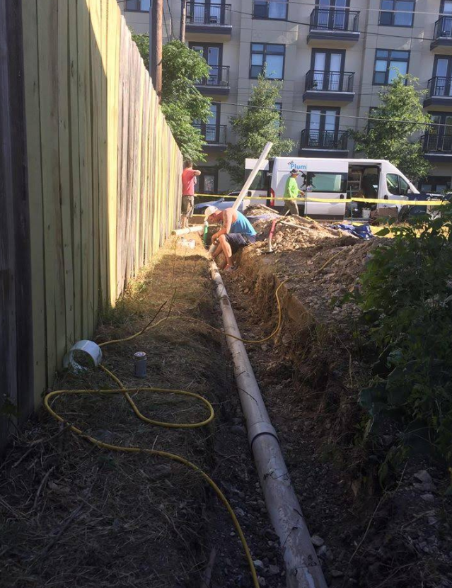 Workers install a pipe in a trench next to a wooden fence with a service van parked in the background.