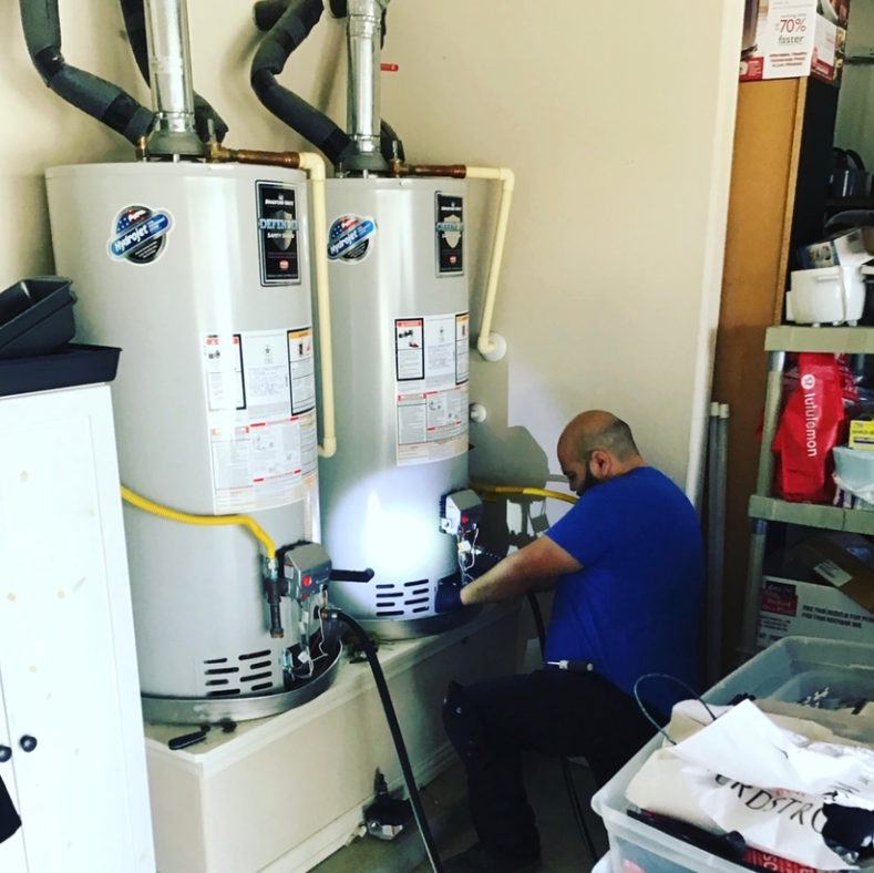 A person in a blue shirt repairs two side-by-side water heaters in a utility room.