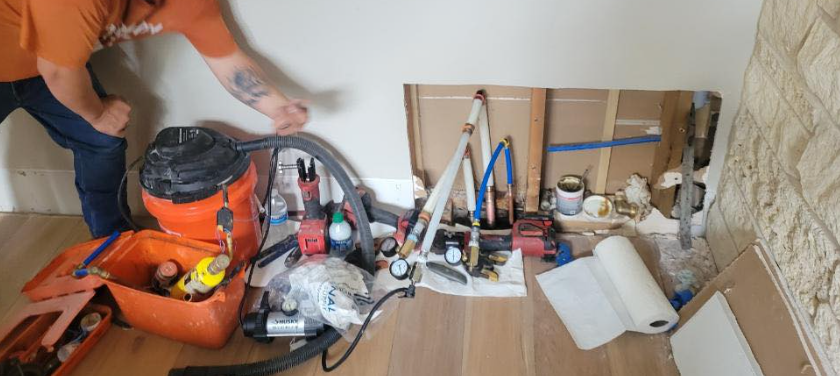 A person in an orange shirt kneels by an open wall cavity containing plumbing pipes and tools, with equipment on the floor.