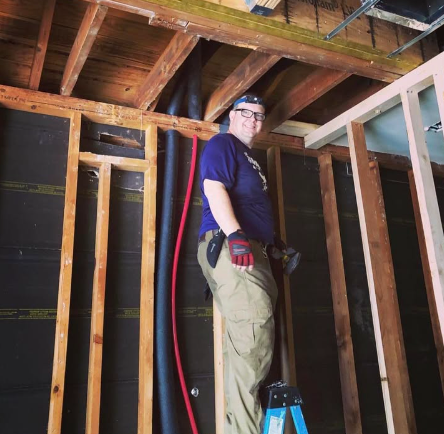 A person wearing work gloves and glasses stands on a blue stepladder while renovating the framing of a wooden wall.