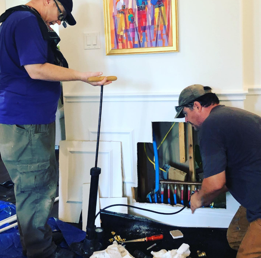 Two workers perform plumbing repairs behind an open wall panel in a home, one using a pump while the other connects pipes.