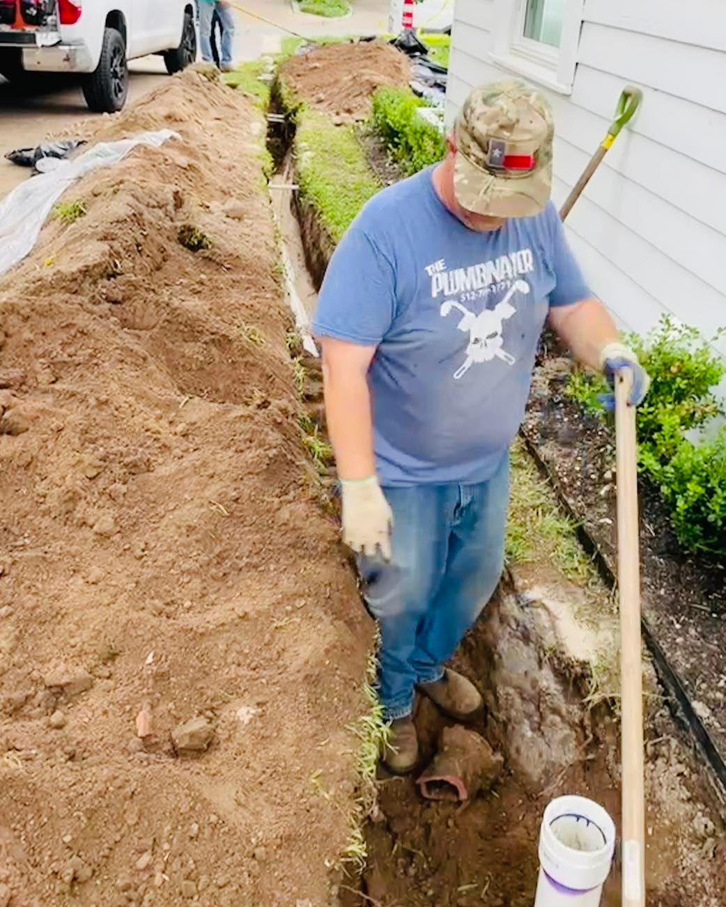 A person wearing a blue hoodie and a light gray baseball cap leans into a dirt-filled hole, working on a utility pipe.