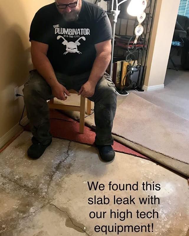 A person wearing a red shirt and cap works on white plastic piping in a dirt-filled crawl space.