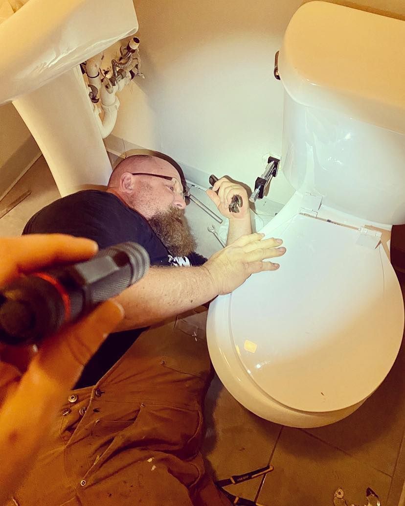 A person kneeling and working on the plumbing under a white farmhouse kitchen sink.