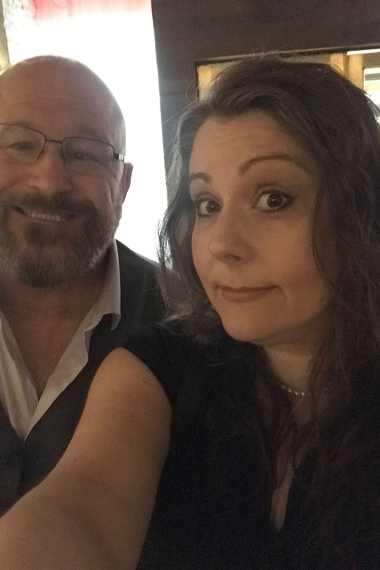 A smiling person with glasses and a beard stands next to a person with long, dark hair in an indoor, lit space.