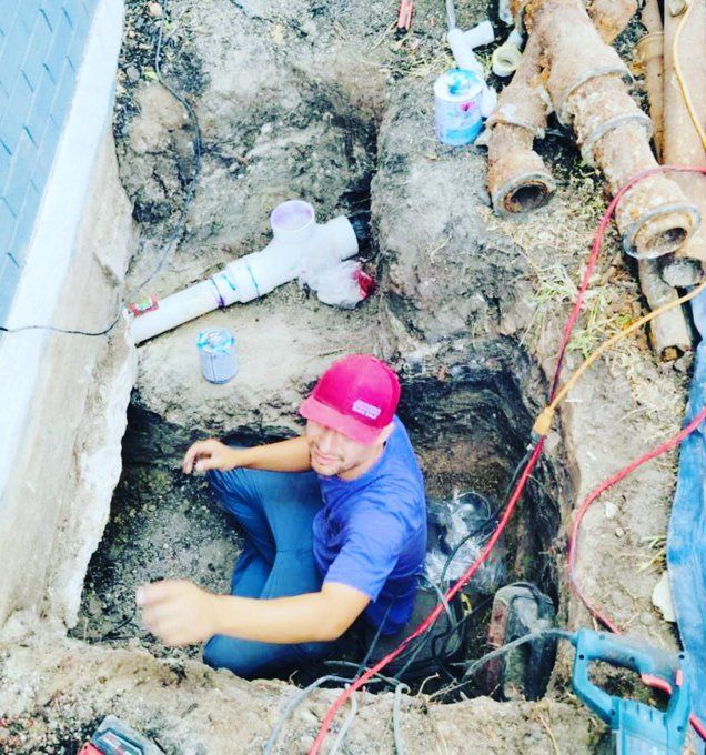 A white PVC pipe runs horizontally through a dirt crawl space, supported by a metal hanger attached to the foundation.