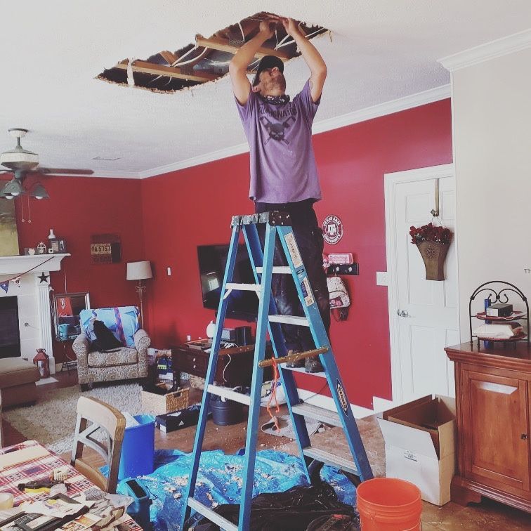 A person in a red shirt and cap works on white PVC piping in a tight, debris-filled crawl space.