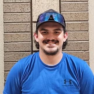 A person smiling while wearing a blue t-shirt, a baseball cap, and blue-tinted sunglasses, standing before a brick wall.