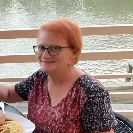 A person with short auburn hair and glasses smiles while sitting at a table with food, outdoors by a water view.