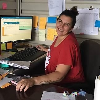A person smiling while sitting at a desk with a computer, surrounded by sticky notes on a wall.