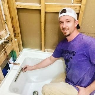 A person in a purple shirt smiles while installing a bathtub drain in a room with exposed wooden wall studs.