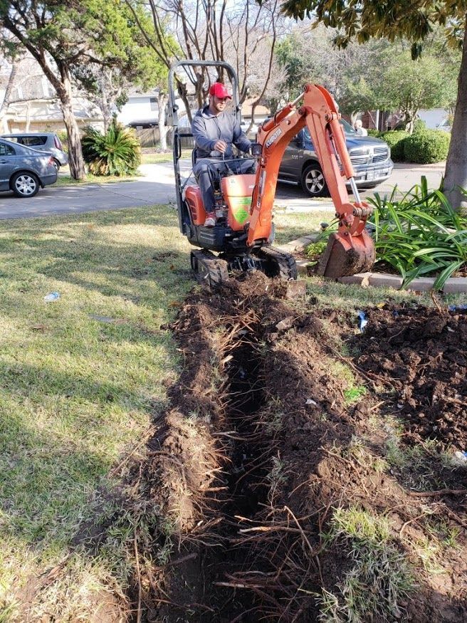 A person in a light green shirt and patterned shorts uses a drain cleaning machine to inspect a pipe in the ground.