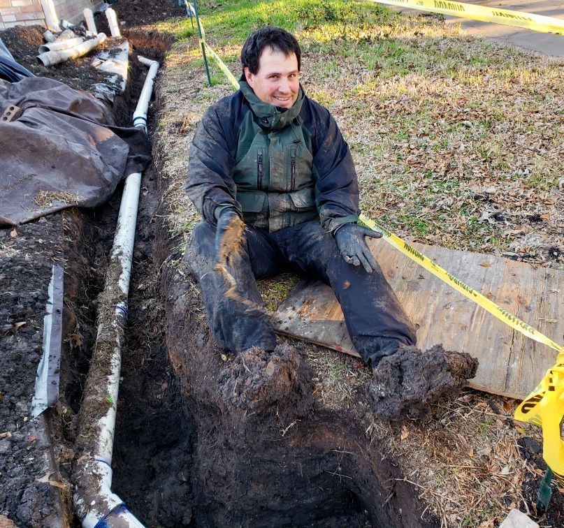 A plumber in a purple shirt works under a kitchen sink, illuminated by a headlamp and a portable work light.