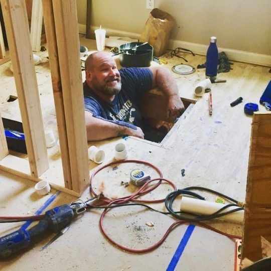 A person wearing a grey hoodie and cap crouches to install plumbing pipes in an unfinished wooden wall.
