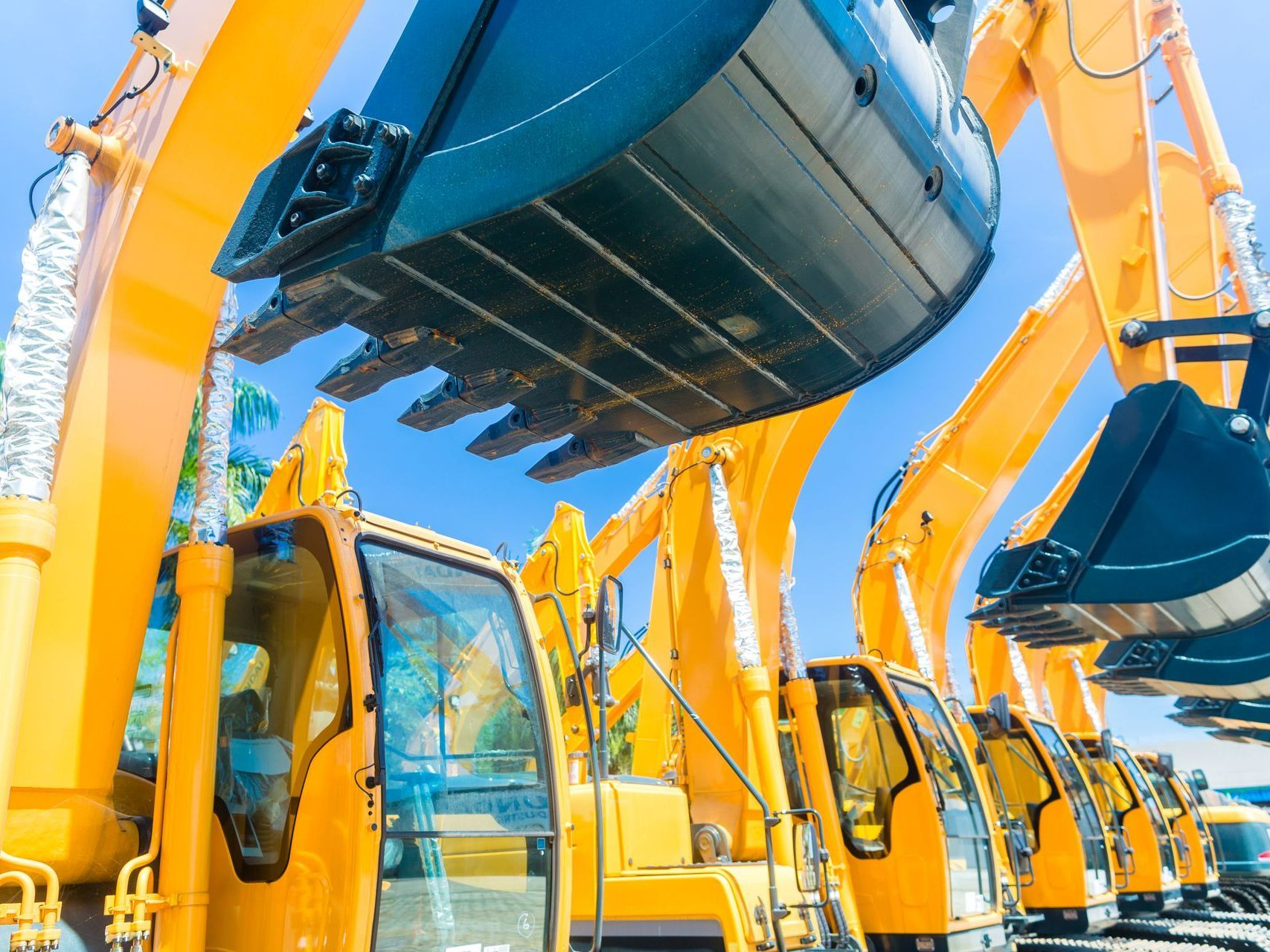 Yellow excavators parked in a row, buckets raised, under a bright blue sky.