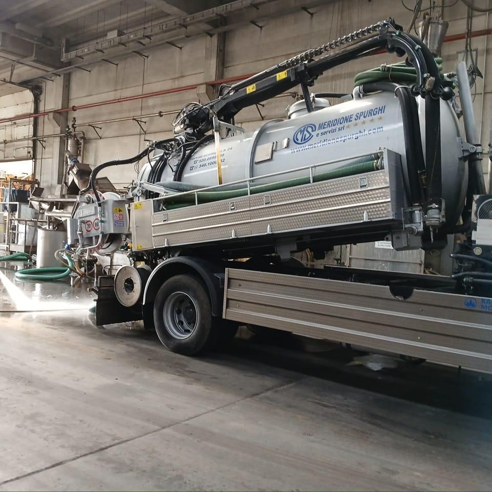 Un camion aspiratore color argento in una fabbrica spruzza acqua, come parte del processo di pulizia.