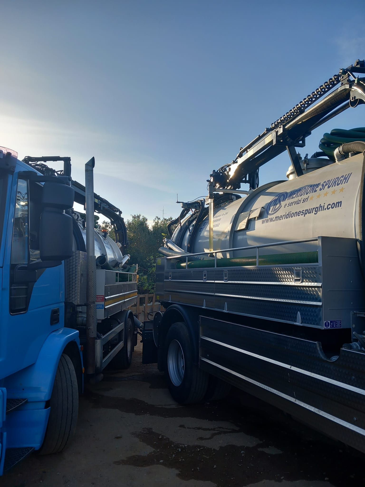 Due autocisterne parcheggiate all'aperto sotto un cielo azzurro, una blu, una nera, con i serbatoi argentati.