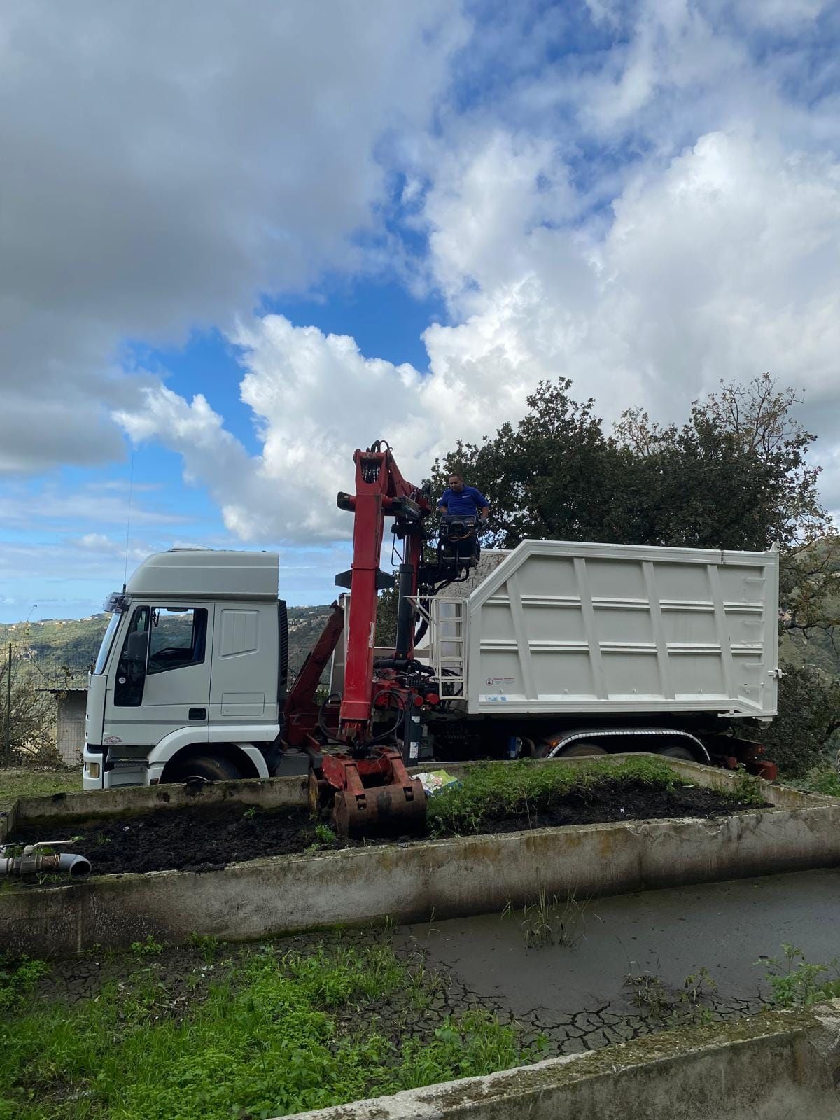 Un camion bianco con un braccio gru rosso carica un container bianco in un ambiente rurale sotto un cielo nuvoloso.