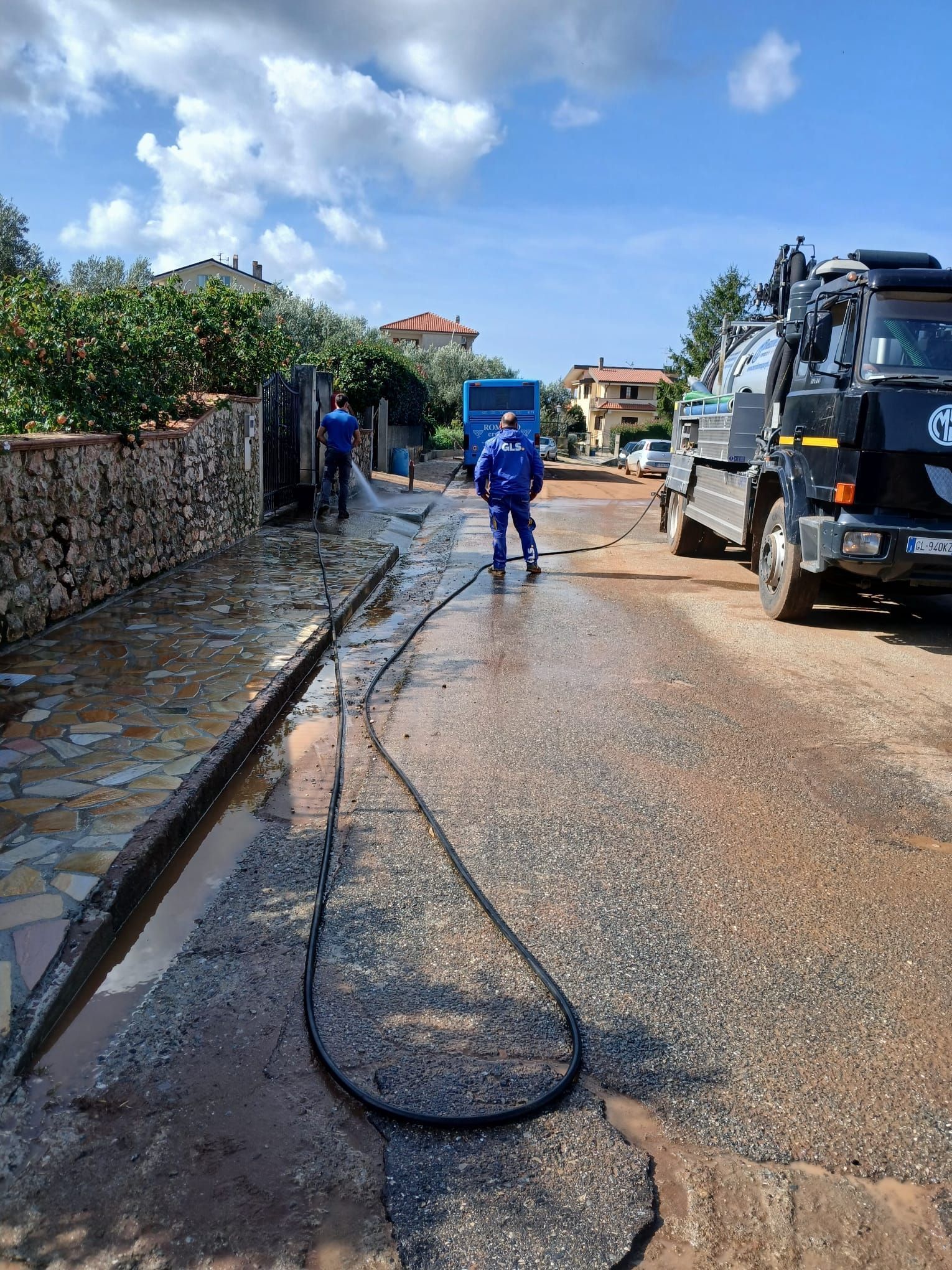 Operai in tuta blu lavano una strada, camion parcheggiato nelle vicinanze, giornata di sole.