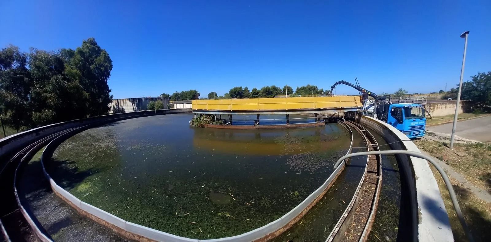 Una piscina circolare per il trattamento delle acque con macchinari e un camion blu, sotto un cielo azzurro e terso.
