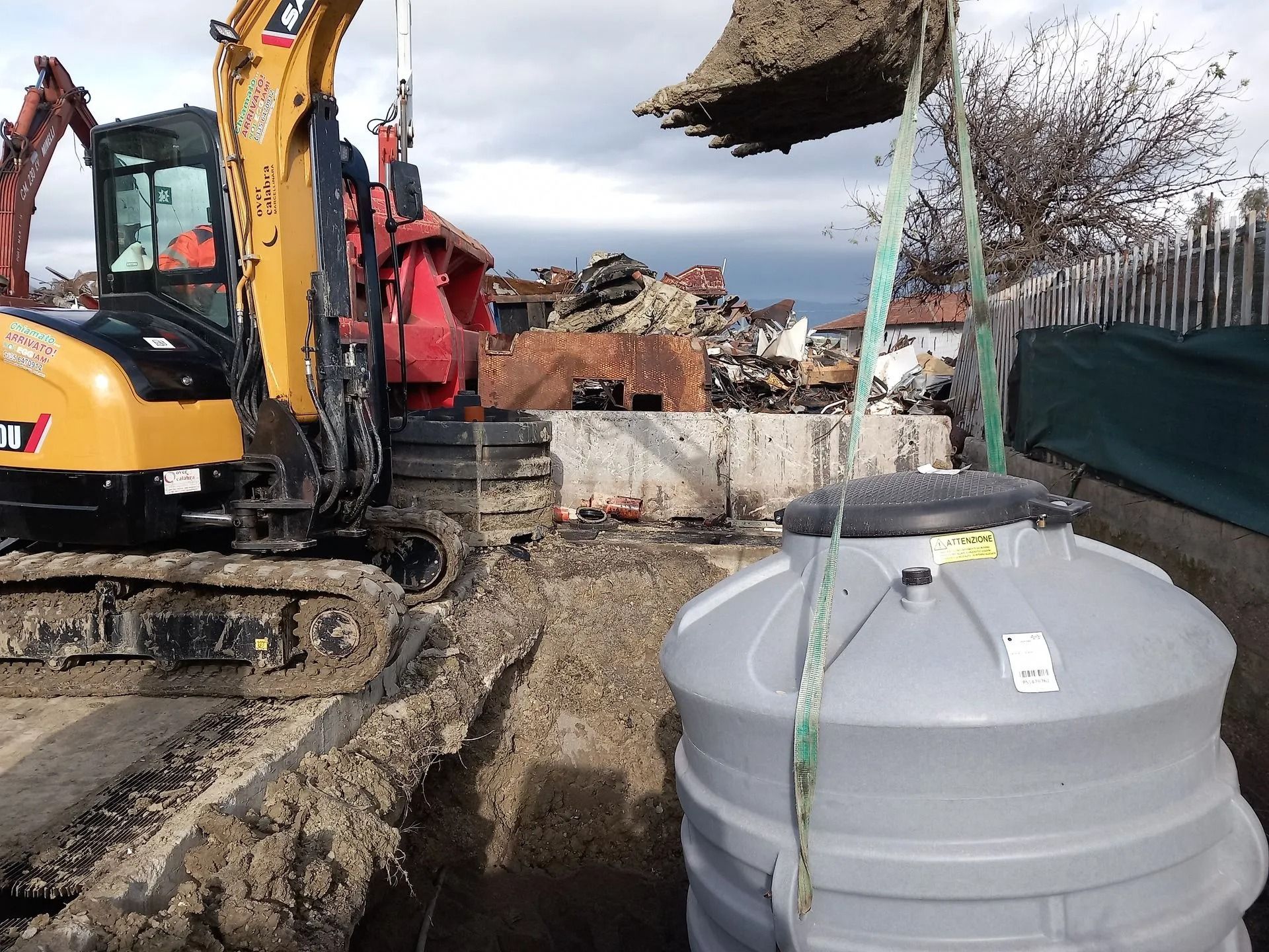 Un escavatore giallo abbassa un serbatoio grigio in una trincea. La terra viene scaricata dalla benna dell'escavatore.
