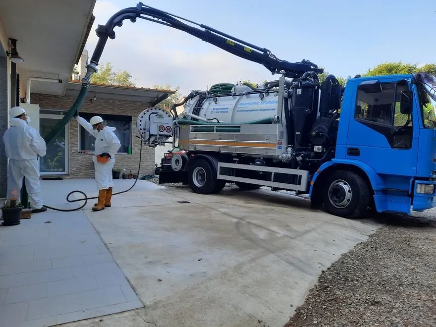 Due persone in tute protettive passano l'aspirapolvere vicino a un edificio con un camion blu e un tubo flessibile.