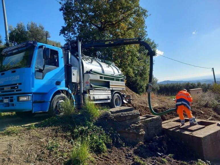 Autocisterna blu che svuota una fossa settica. Operaio con giubbotto arancione guida il tubo. Scenario esterno soleggiato.