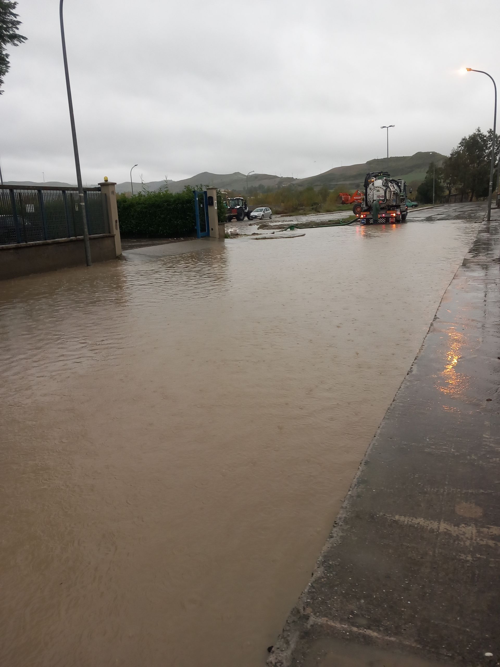 Strada allagata con acqua marrone torbida; escavatore e persone visibili; giornata nuvolosa.