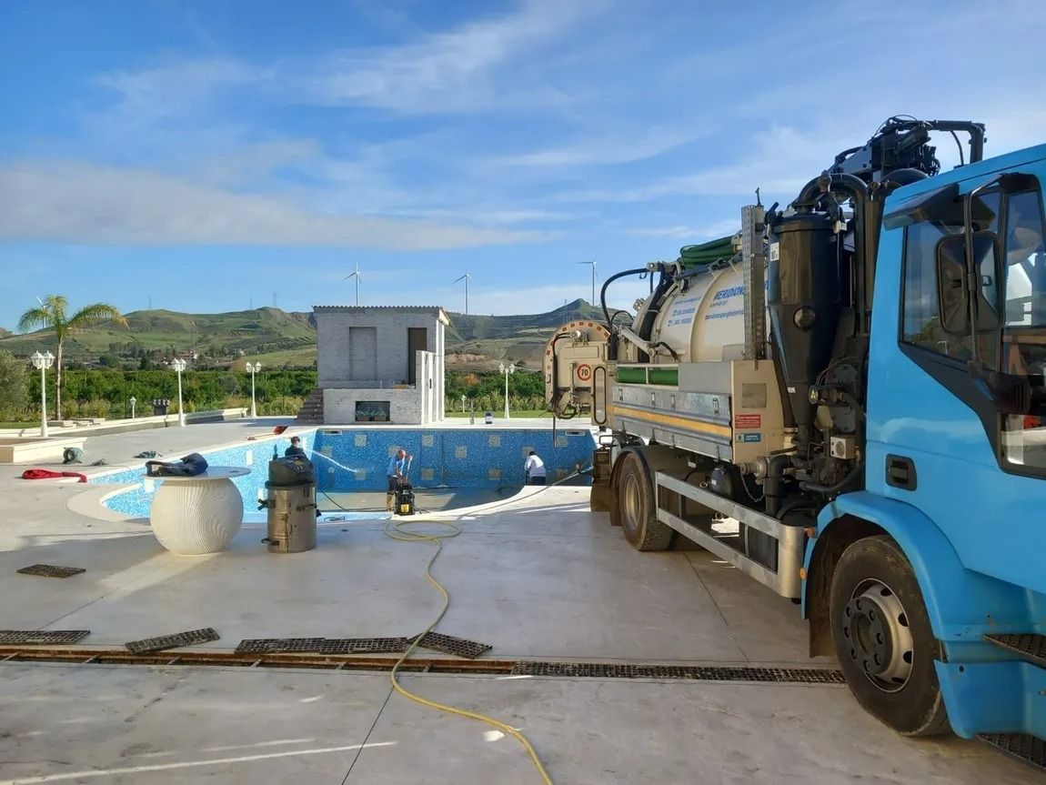Un camion blu aspira una piscina svuotata con un piccolo edificio sullo sfondo sotto un cielo azzurro.