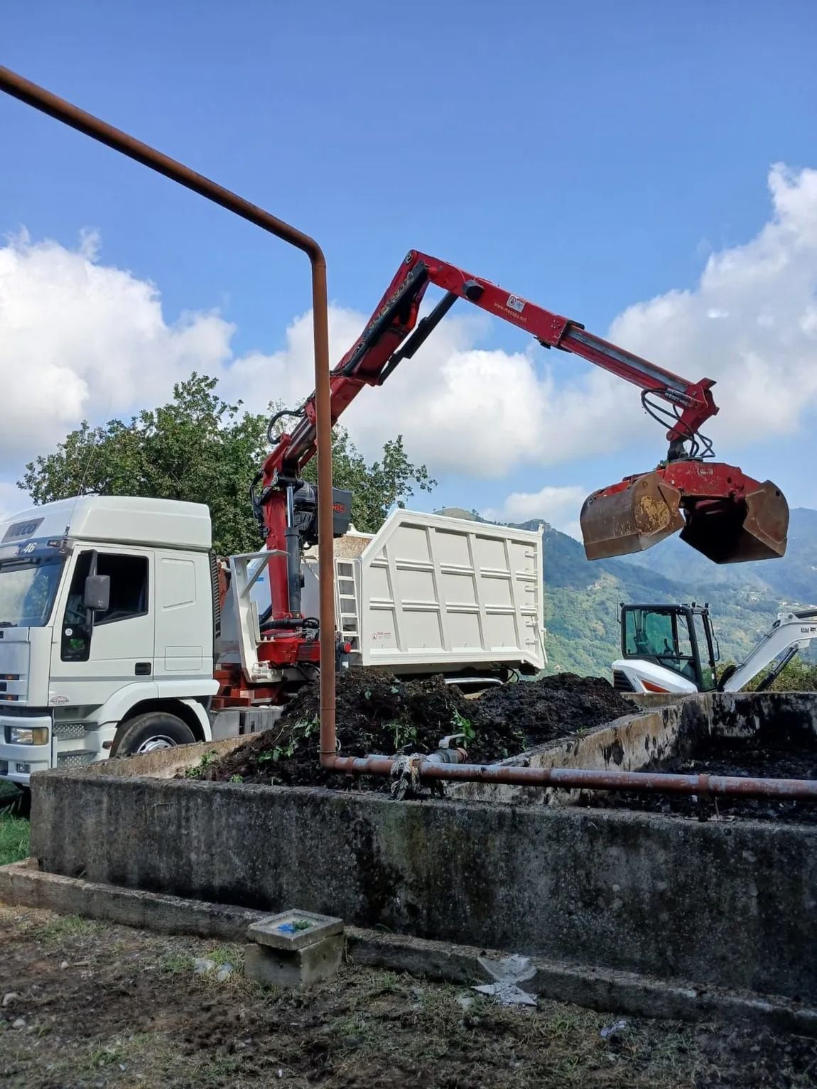Una gru rossa carica la terra da un mucchio su un camion bianco in una giornata di sole, con le montagne sullo sfondo.
