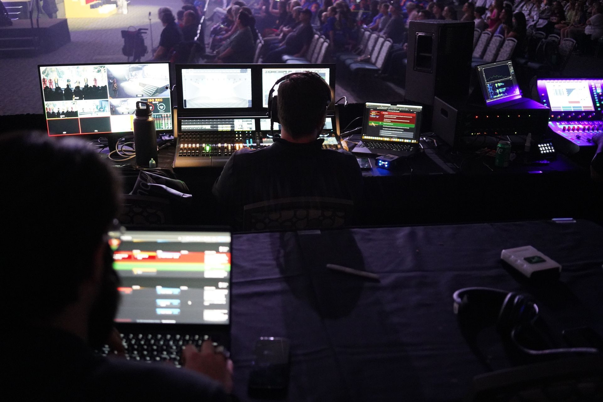 Two people operate video equipment with monitors in a dimly lit room, audience in background.