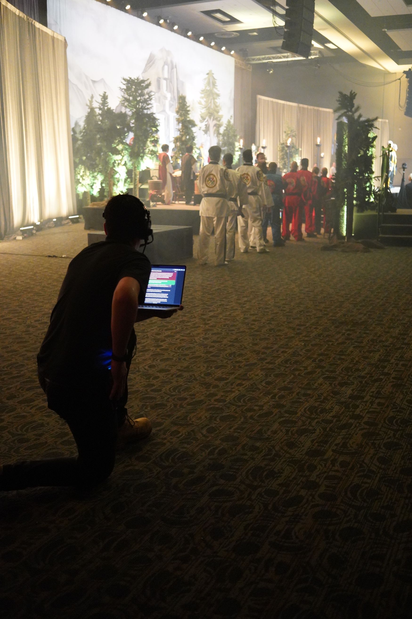 Person kneels, holding a laptop, filming costumed performers on a stage with a mountain backdrop.