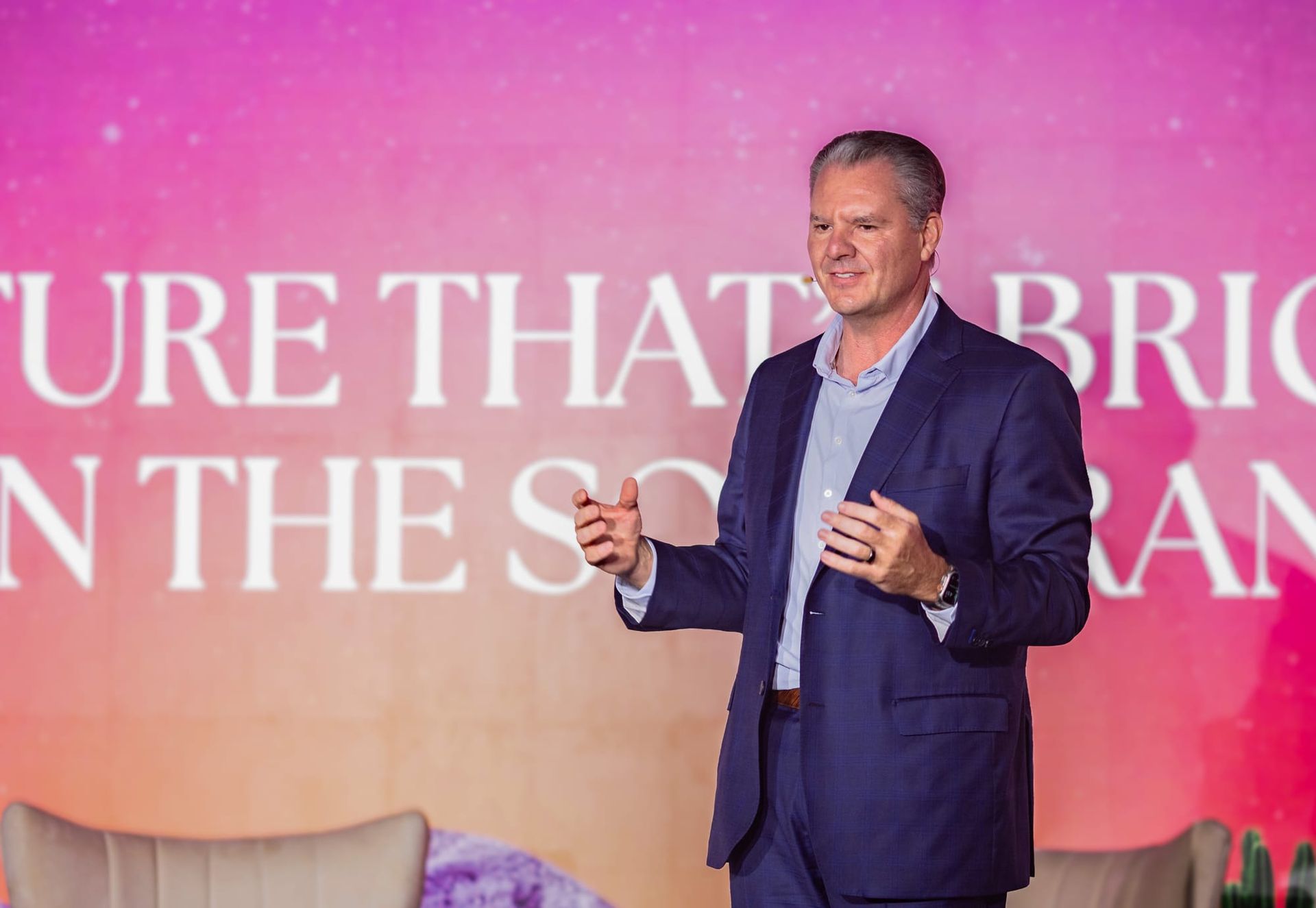 Man in blue suit giving a presentation with hands raised, on a stage with pink and white background.