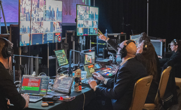 Control room: People at computers and monitors, directing a live broadcast. Some wear headsets, pointing and working.