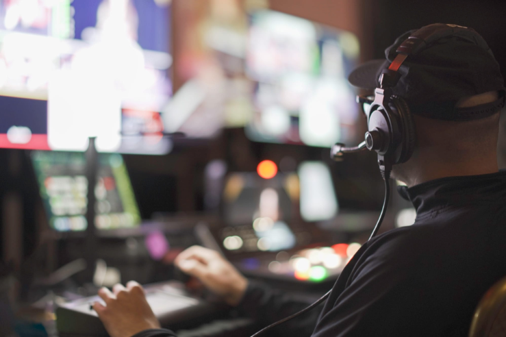 Man with headset operates video equipment in a control room with multiple monitors.