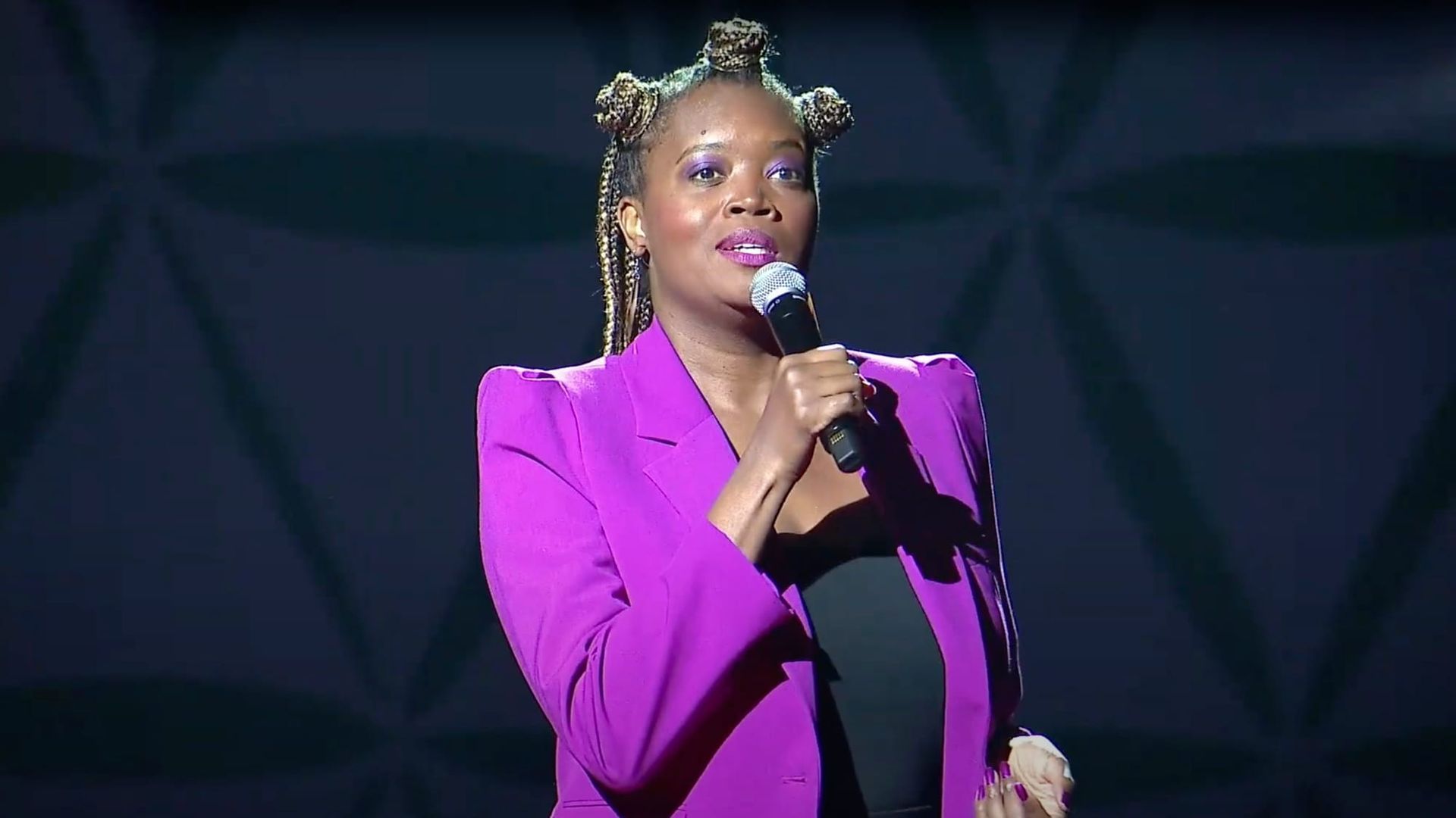 Woman in pink blazer on stage, holding microphone, speaking.