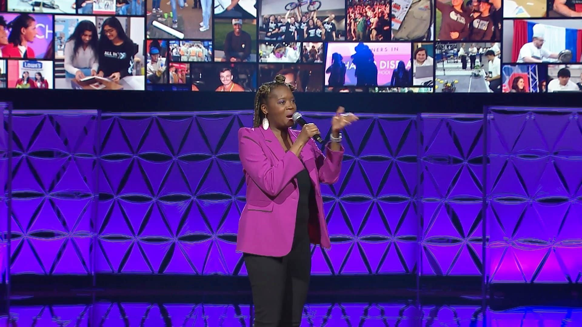 Woman in pink blazer speaking on stage with a screen showing photos in the background.