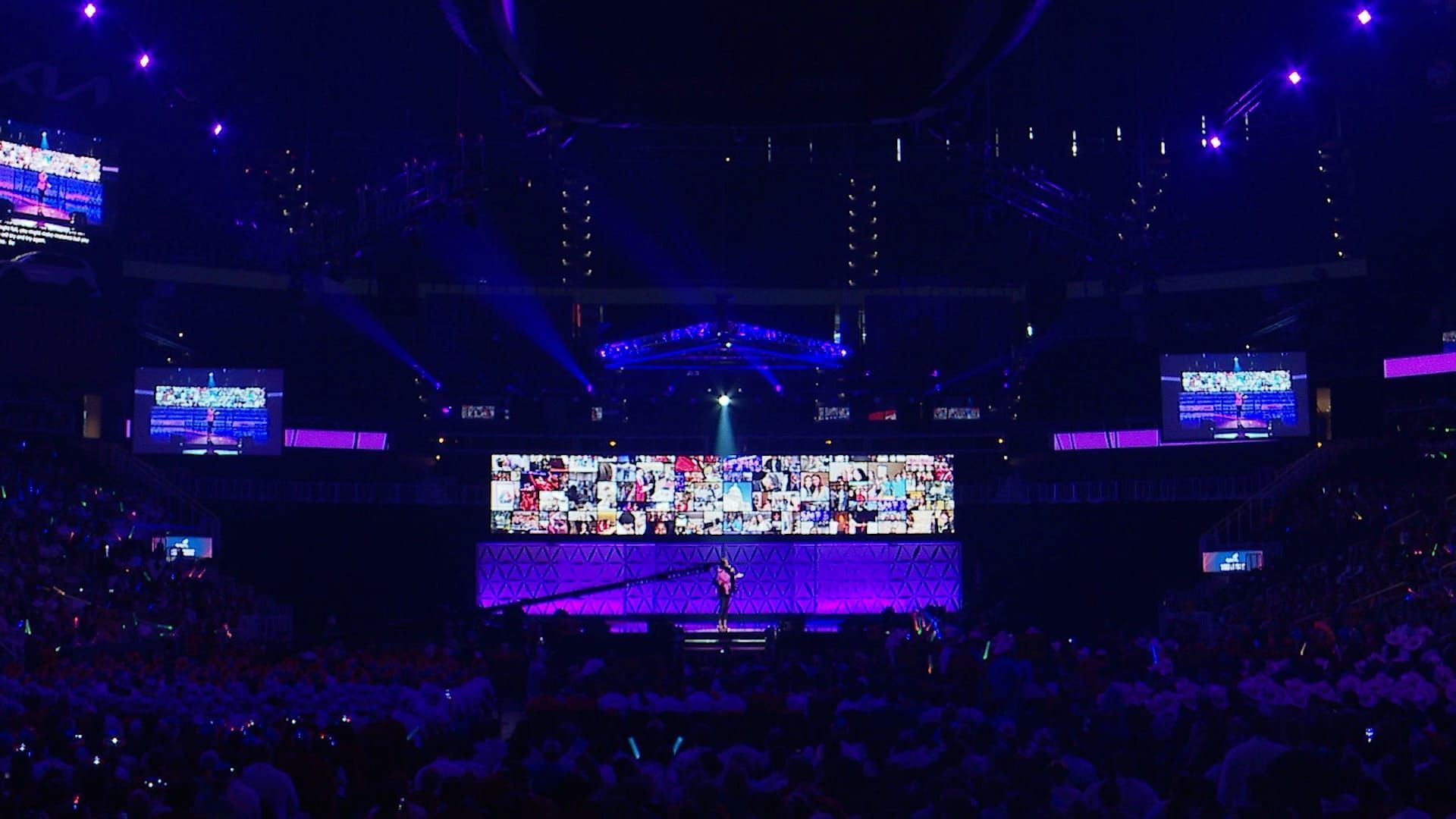Concert stage with large screen displaying images. Audience with raised arms in a dark arena lit by purple lights.