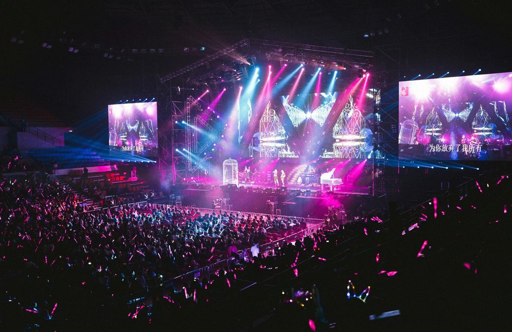 Concert stage with bright pink and blue lights, crowd holding illuminated sticks.
