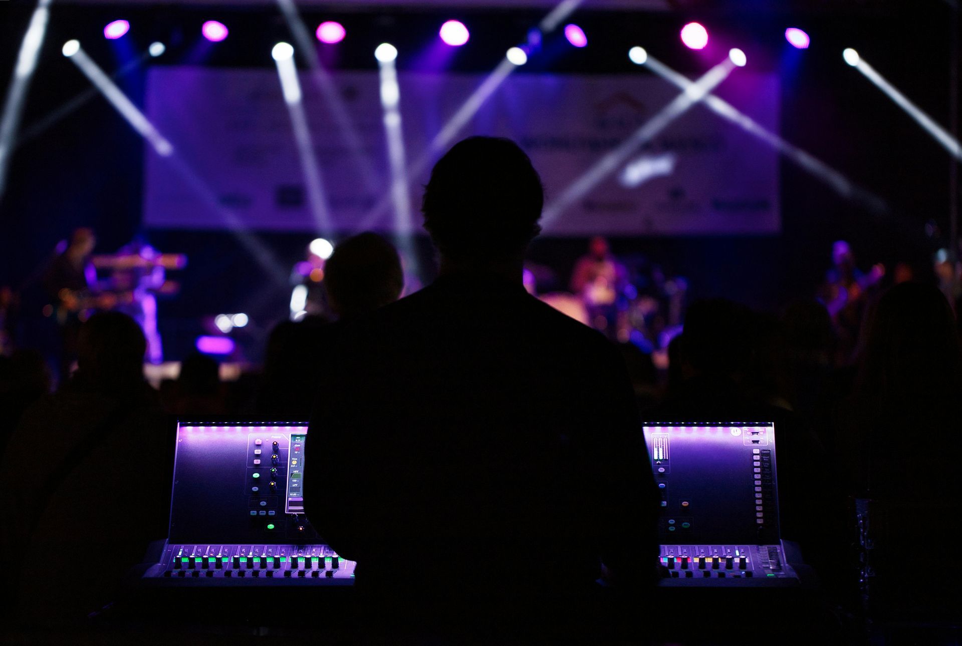 Silhouette of a sound engineer at a mixing board, with a band performing on stage lit in purple.
