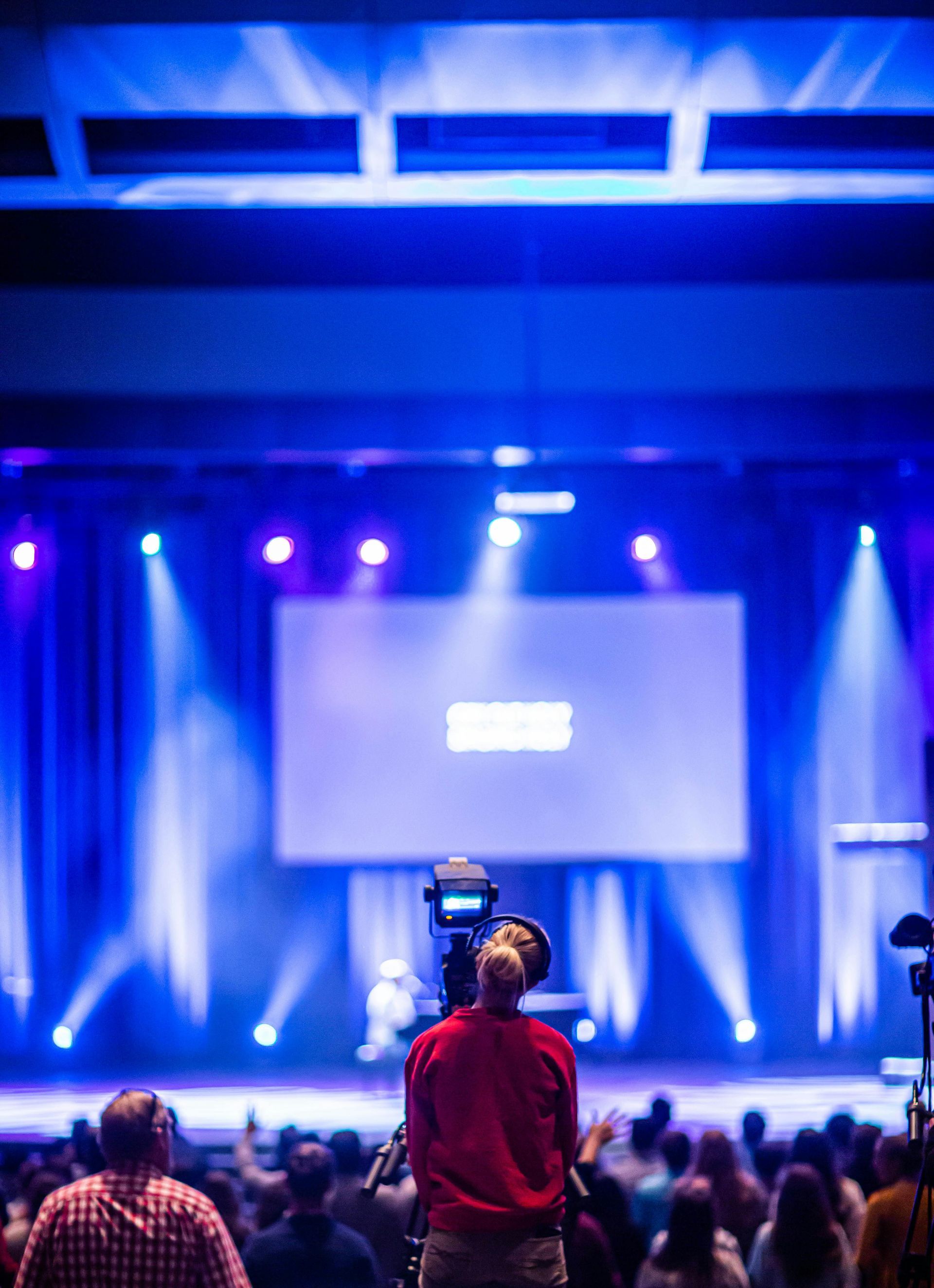 Person filming a presentation on stage with blue lights and a large screen; audience in the foreground.
