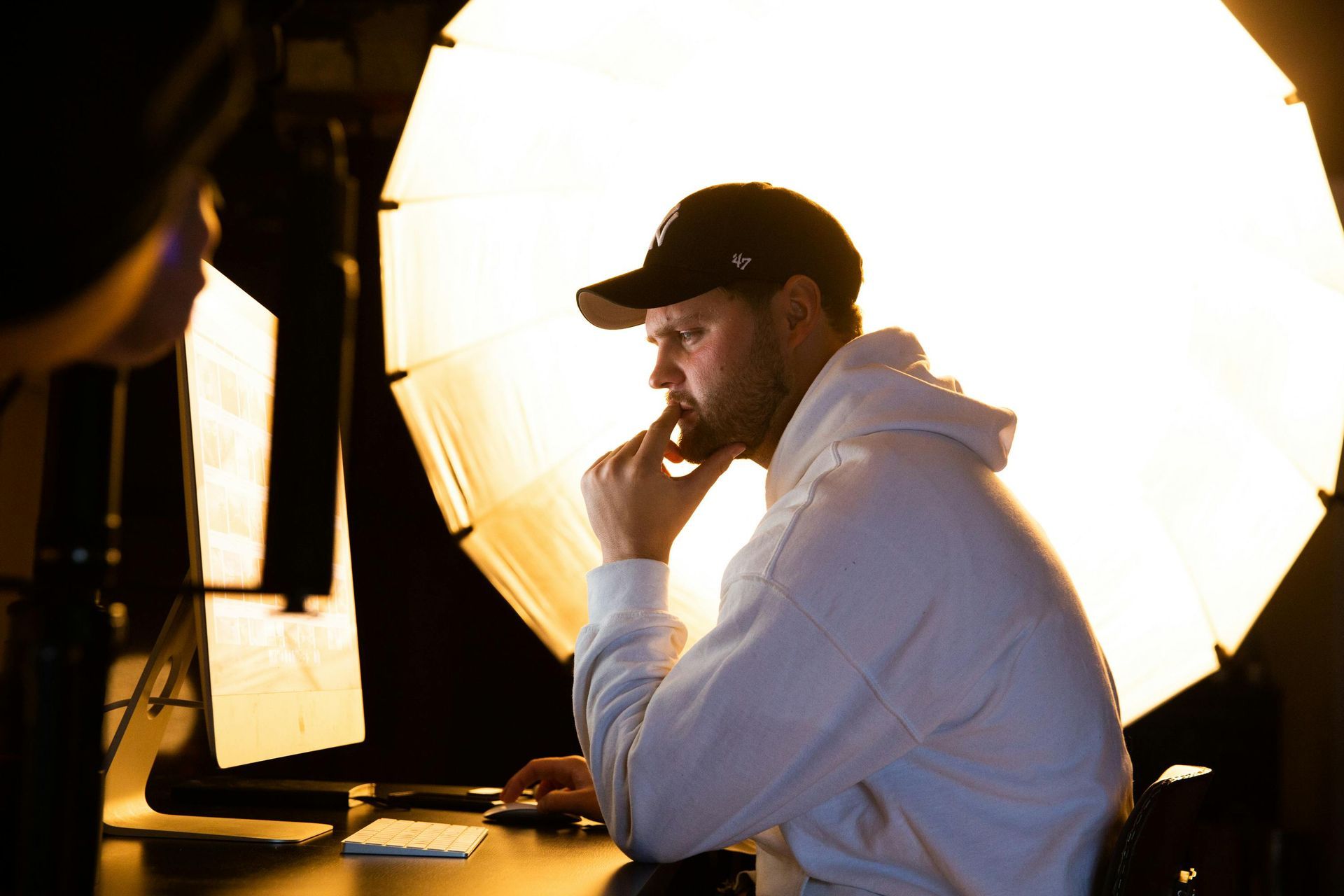 Man in a hoodie and baseball cap, illuminated by a bright light, appears thoughtful, working on a computer.