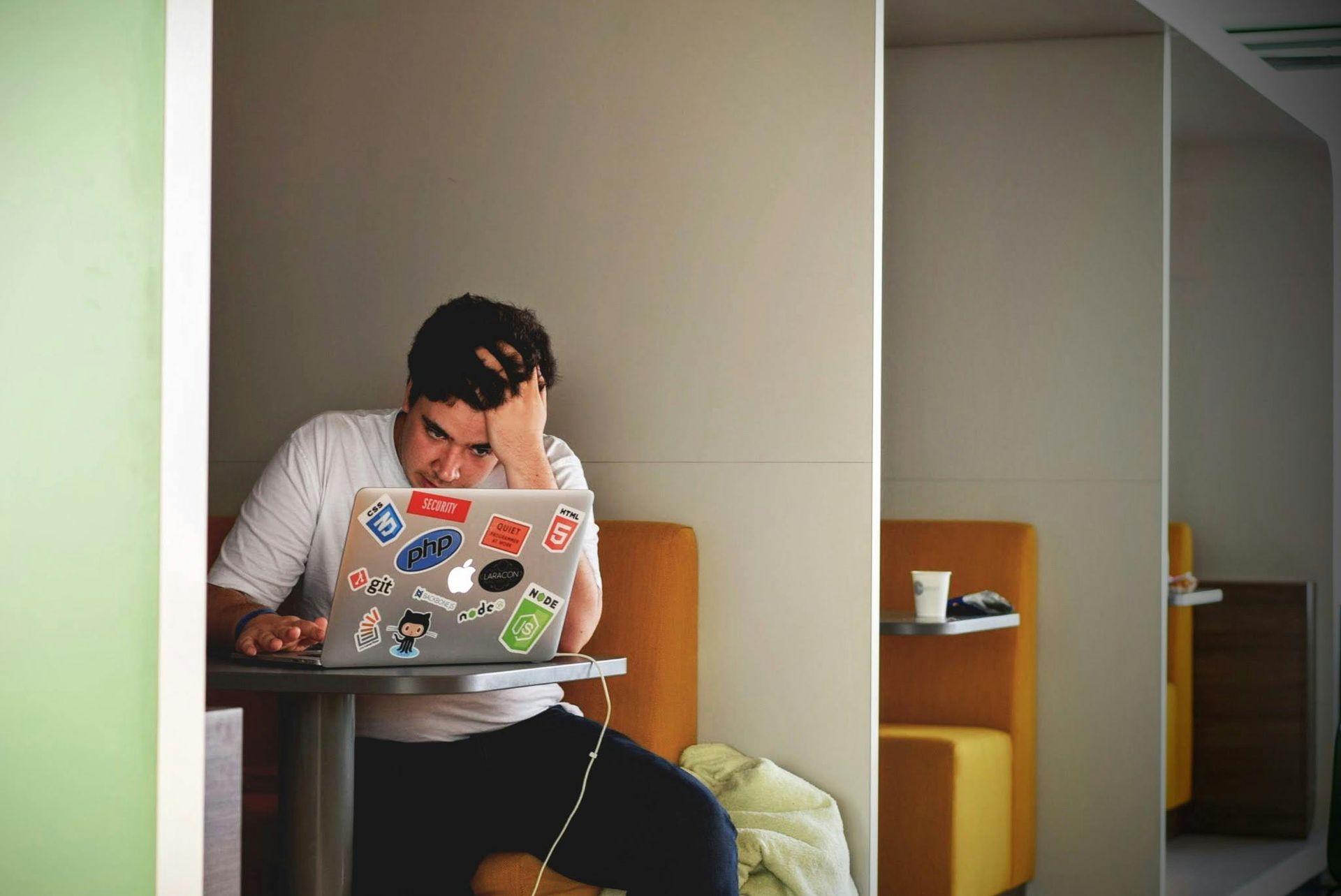 Person with hand on head, stressed, working on laptop in a small booth, with a white cup.