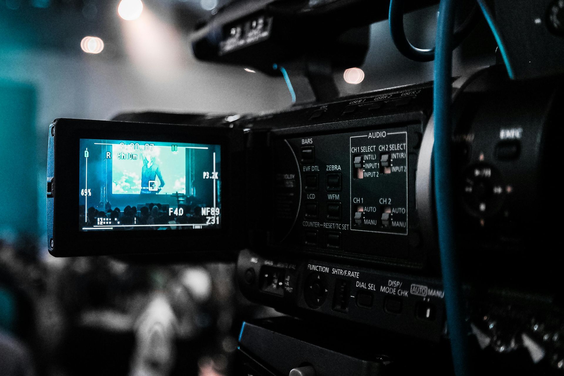Close-up of a video camera filming a brightly lit stage. The screen displays a person performing.