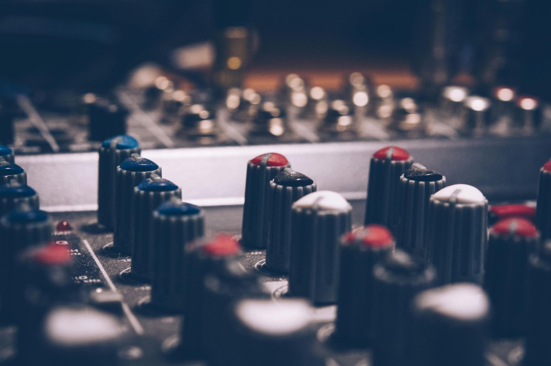 Close-up of a sound mixing console with numerous knobs in various colors, primarily blue, red, and white.