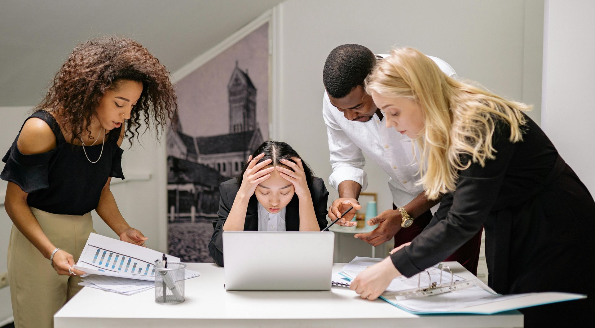 Four office workers around a table, one woman stressed, two looking at papers, and one pointing.