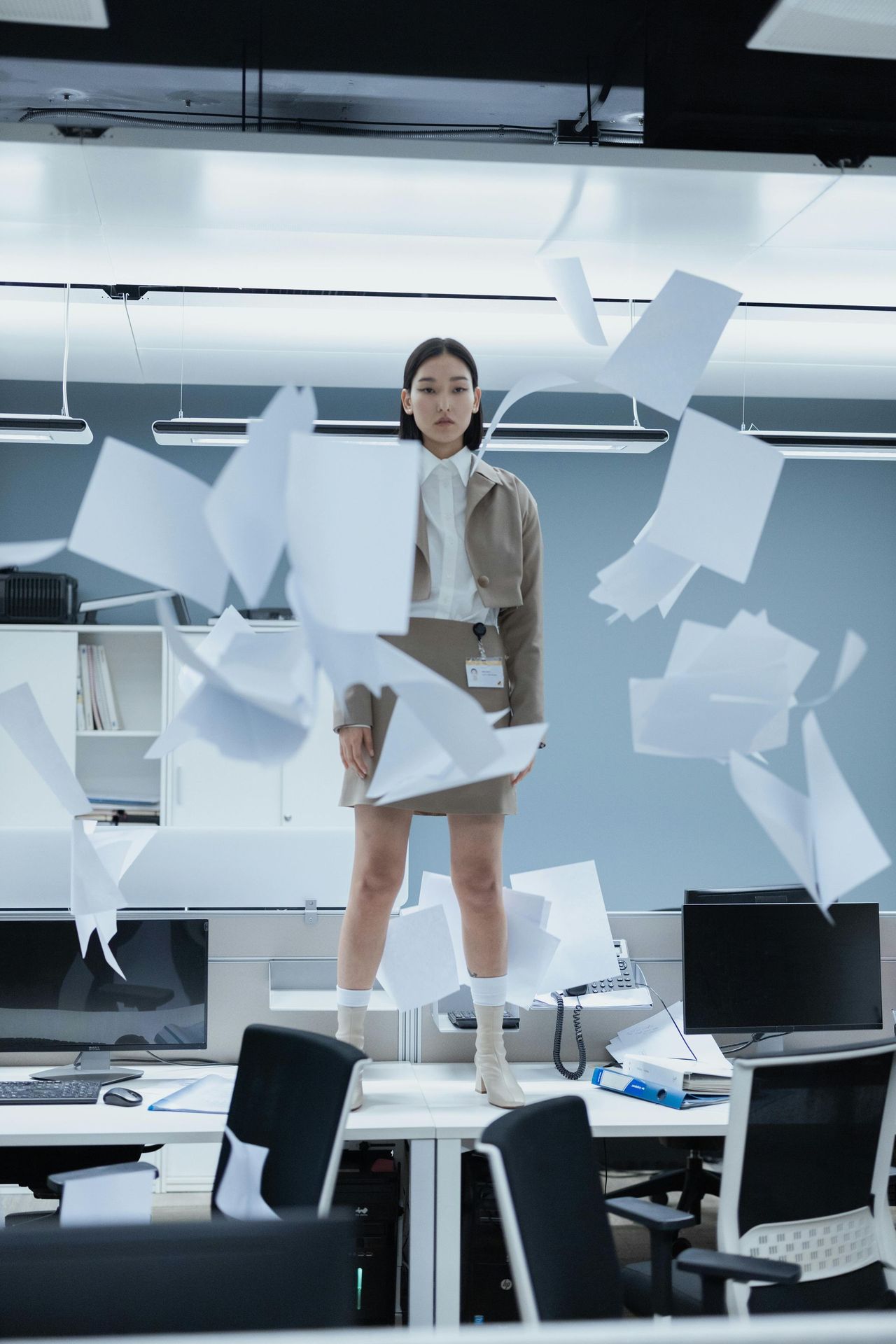 Woman standing on office desk, papers flying. Beige outfit, office setting, determined expression.