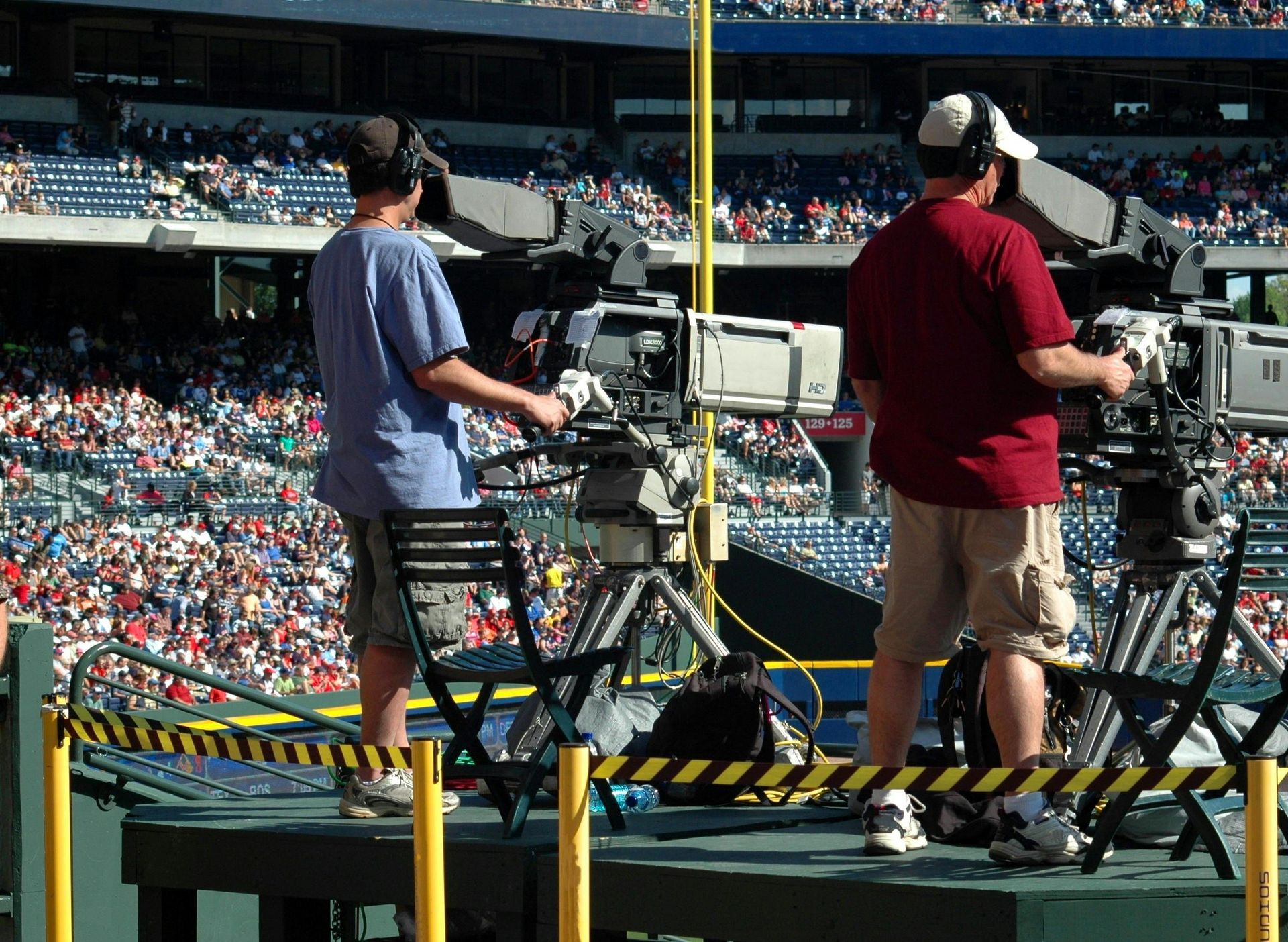 Two people operating cameras at a baseball stadium, filming the game with fans in the background.